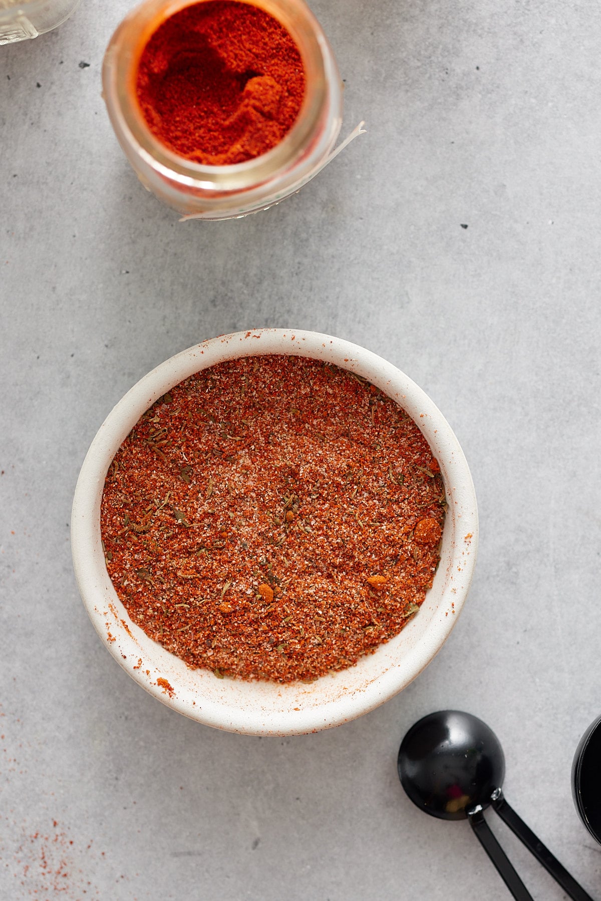 A white bowl containing a blend of spices, with an open jar of paprika and some measuring spoons set alongside