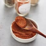 White bowl and a wooden spoon with spice blend and jars of herbs and spices set alongside