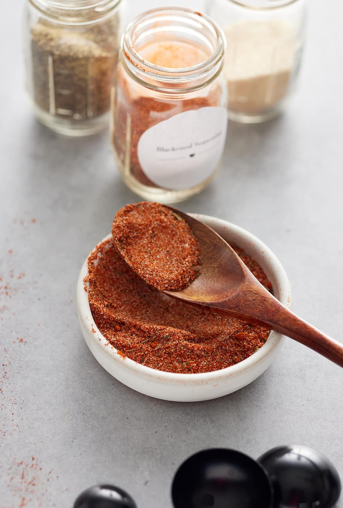 White bowl and a wooden spoon with spice blend and jars of herbs and spices set alongside