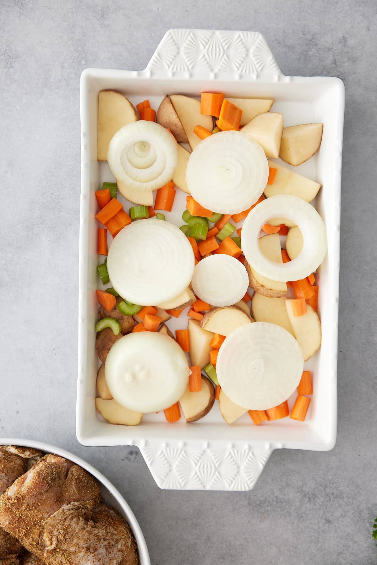 White oven proof dish containing layer of vegetables including potato carrots, celery and onion