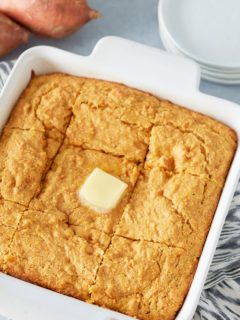 White square oven proof dish with baked sweet potato cornbread and square of melted butter on top and serving plates and whole sweet potatoes set alongside