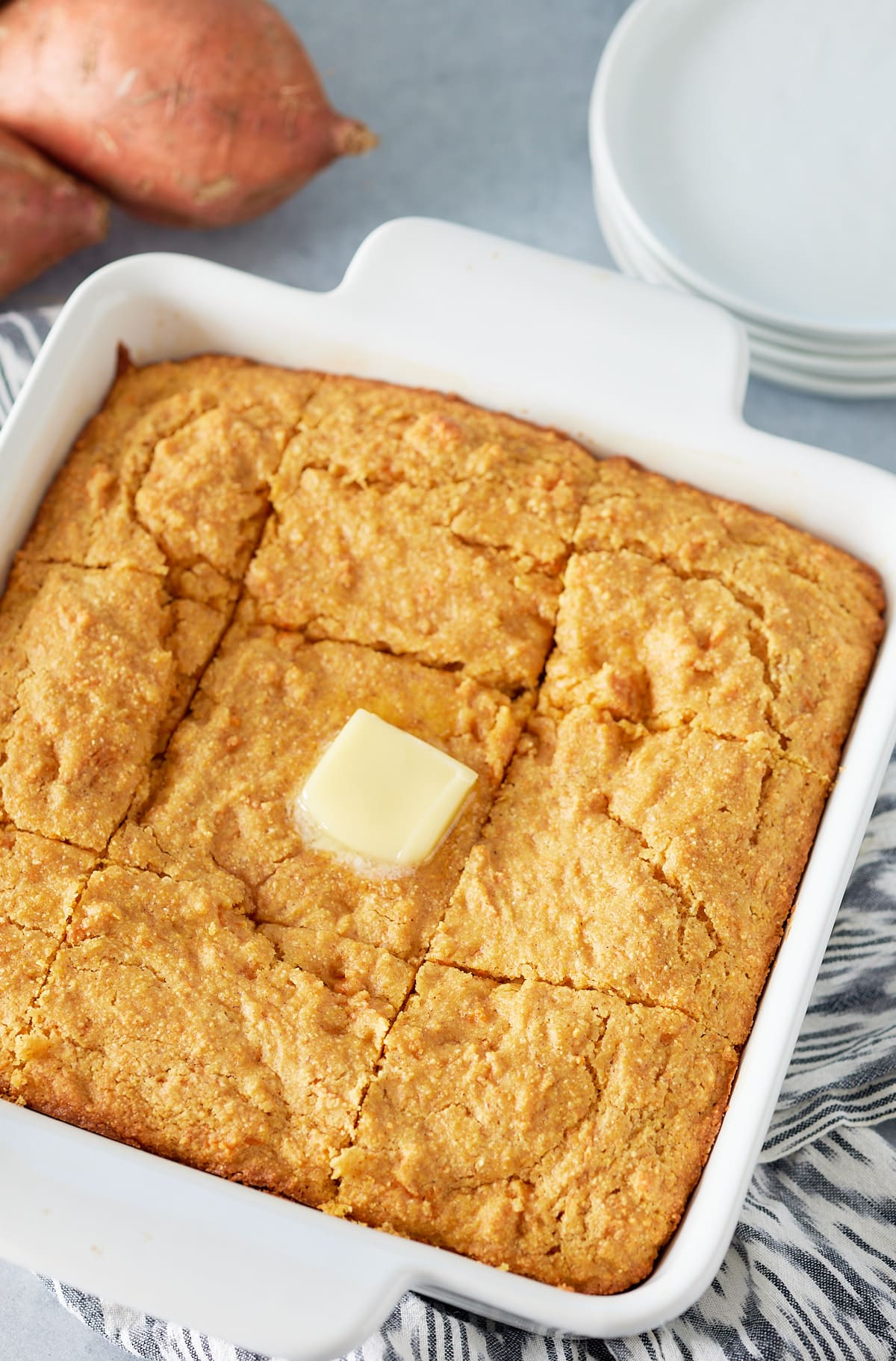 White square oven proof dish with baked sweet potato cornbread and square of melted butter on top and serving plates and whole sweet potatoes set alongside