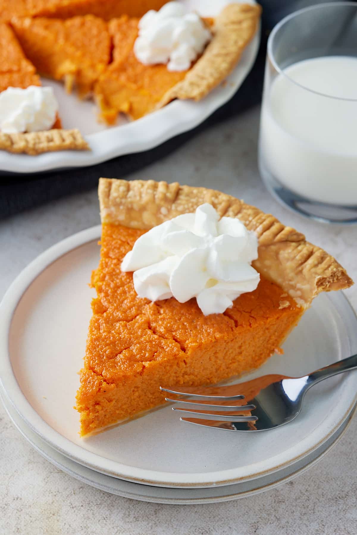 A white plate with a slice of carrot pie topped with piped cream with the whole pie and a glass of milk set alongside