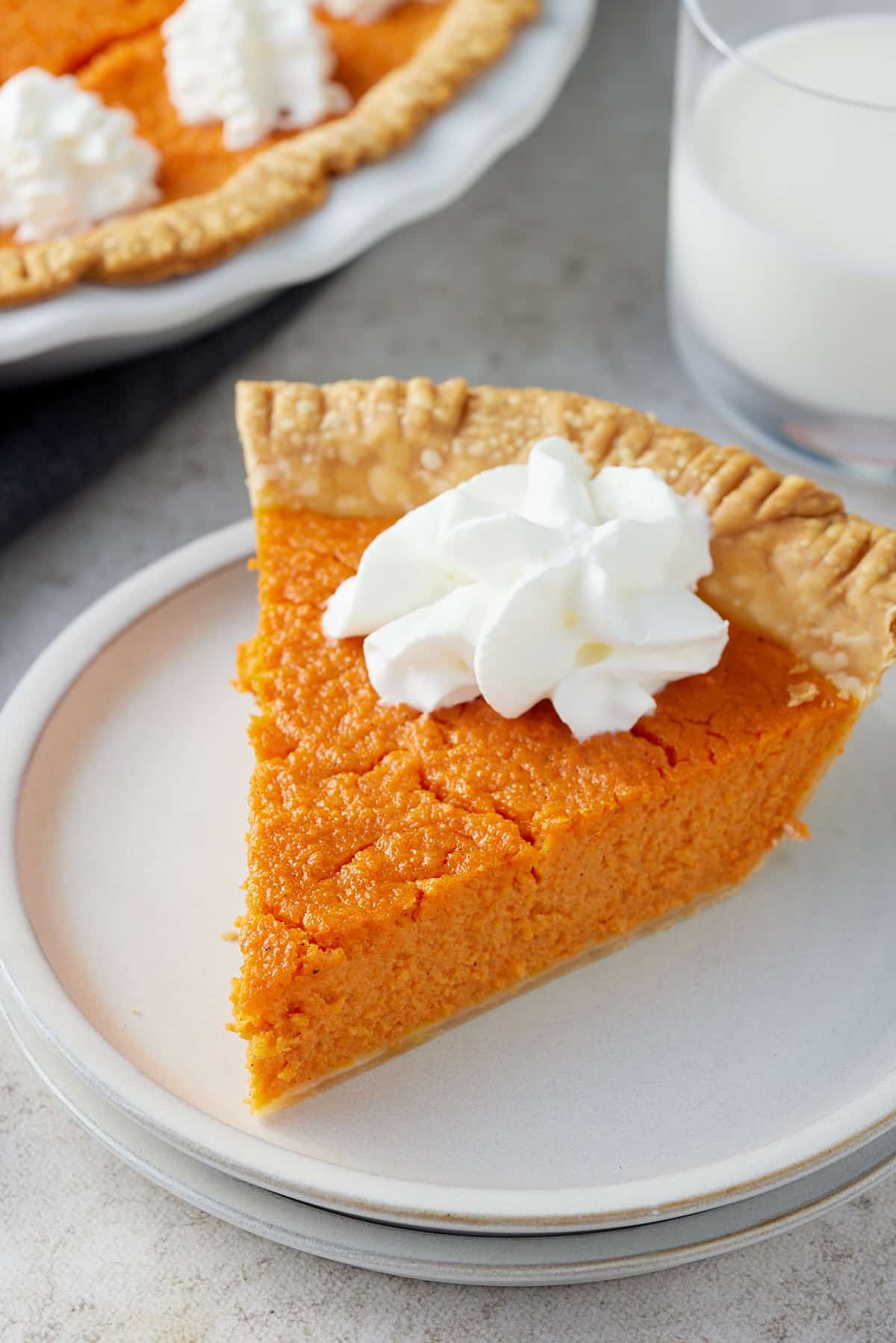A white plate with a slice of carrot pie topped with piped cream with the whole pie and a glass of milk set alongside