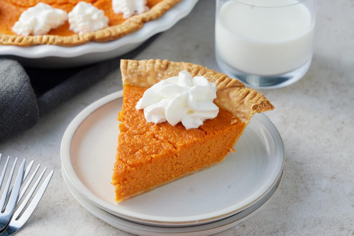 A white plate with a slice of carrot pie topped with piped cream with the whole pie and a glass of milk set alongside