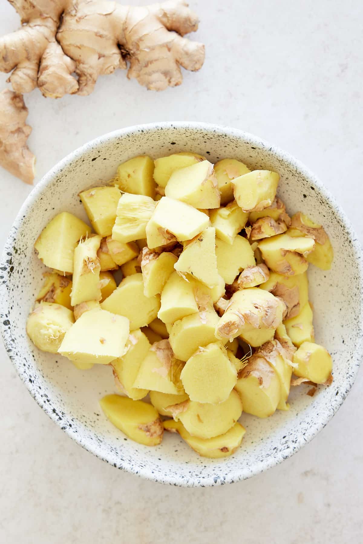 A white bowl of roughly chopped ginger root with whole root of ginger set alongside