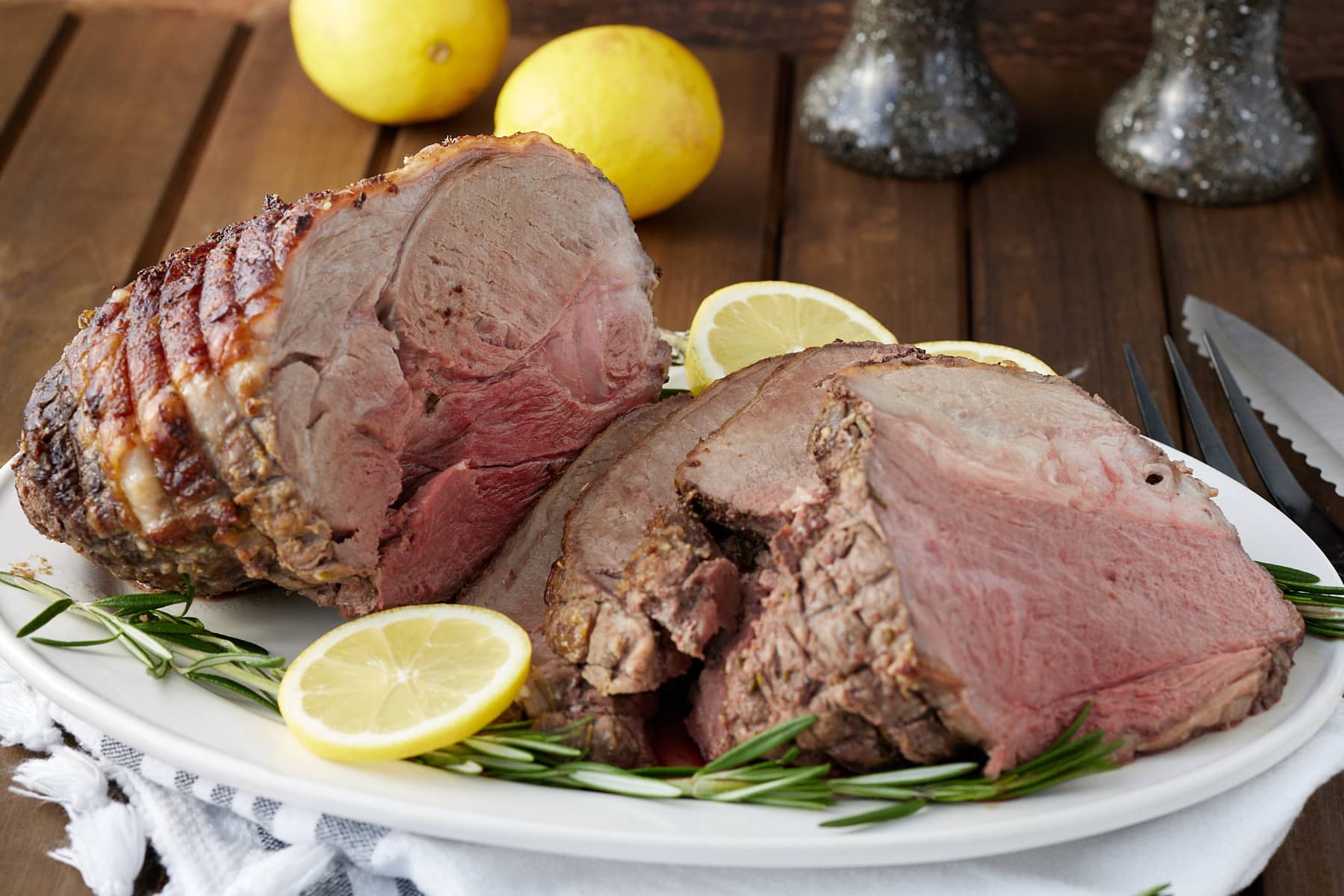 Whole roasted boneless leg of lamb sliced on a serving platter with fresh lemon and rosemary garnish