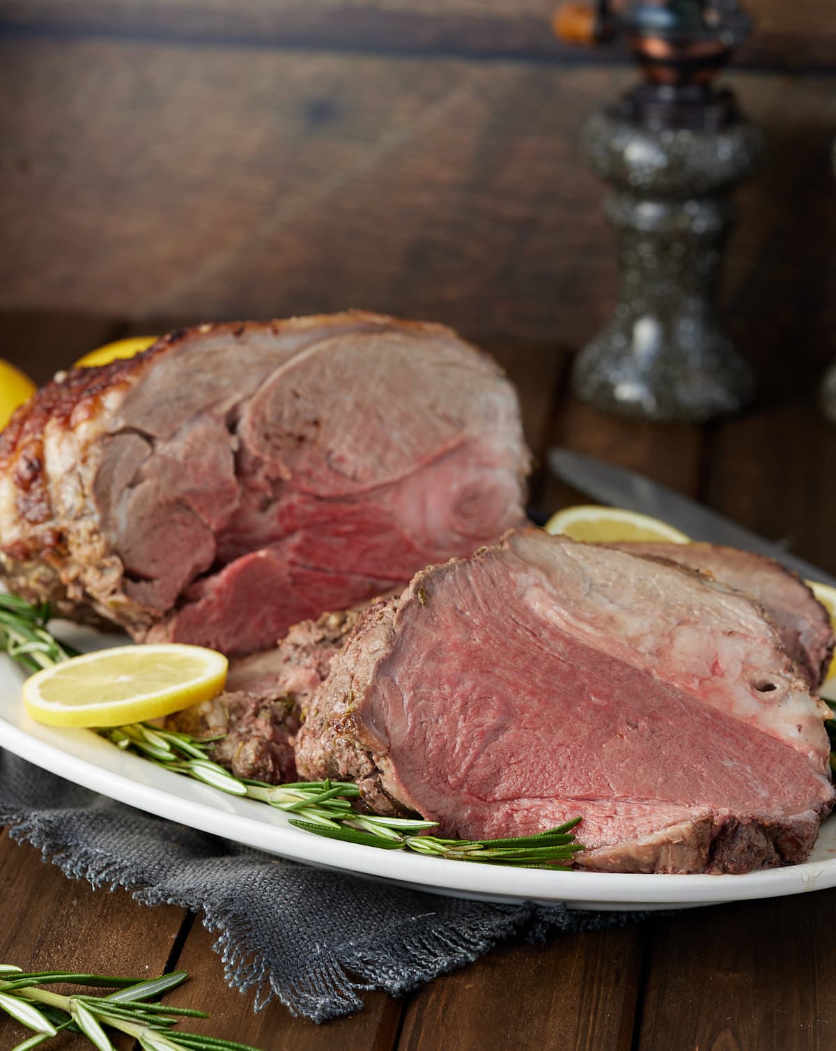 Whole roasted boneless leg of lamb sliced on a serving platter with fresh lemon and rosemary garnish