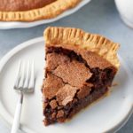 A slice of chocolate chess pie on a plate with a serving fork and the larger pie set alongside