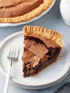 A slice of chocolate chess pie on a plate with a serving fork and the larger pie set alongside