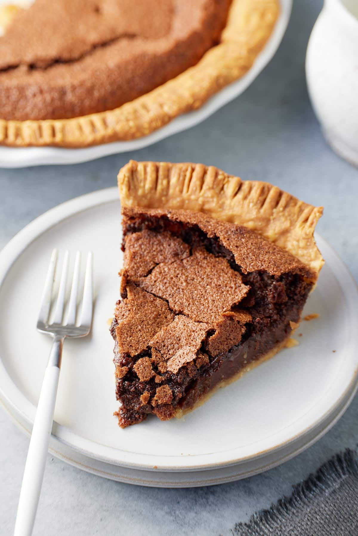 A slice of chocolate chess pie on a plate with a serving fork and the larger pie set alongside