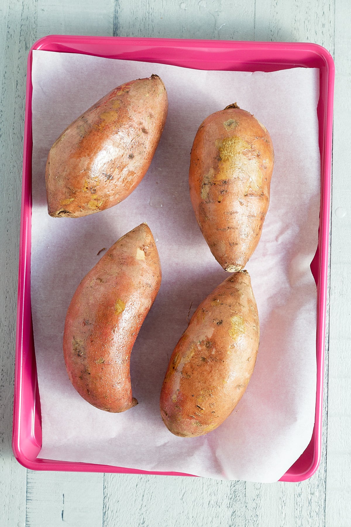 four sweet potatoes on a pink baking tray lined with parchment paper