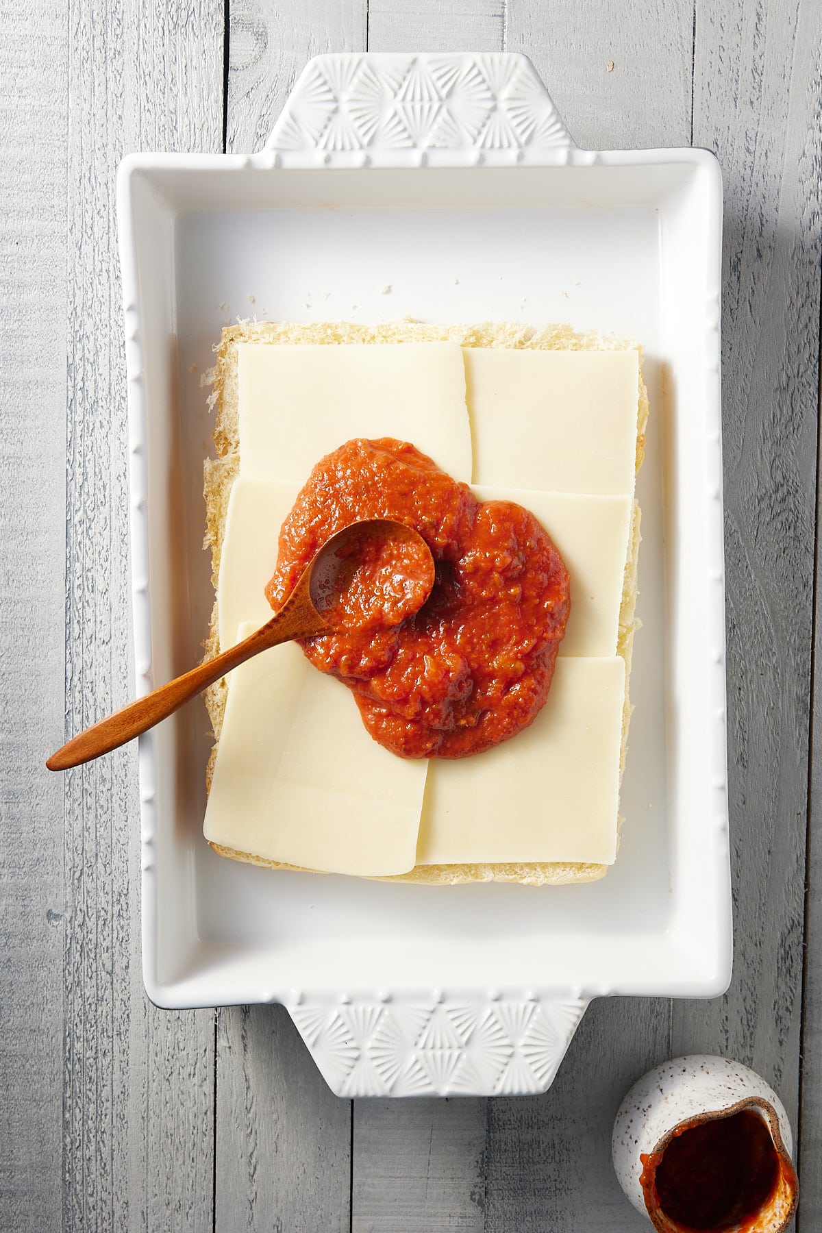 white oven proof dish with base of bread rolls topped with sliced mozzarella cheese and a spoon of pizza sauce