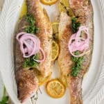 white serving platter with 2 cooked whole fish stuffed with fresh thyme and lemon slices and garnished with red onion and a drizzle of olive oil