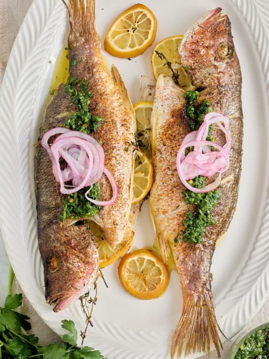white serving platter with 2 cooked whole fish stuffed with fresh thyme and lemon slices and garnished with red onion and a drizzle of olive oil