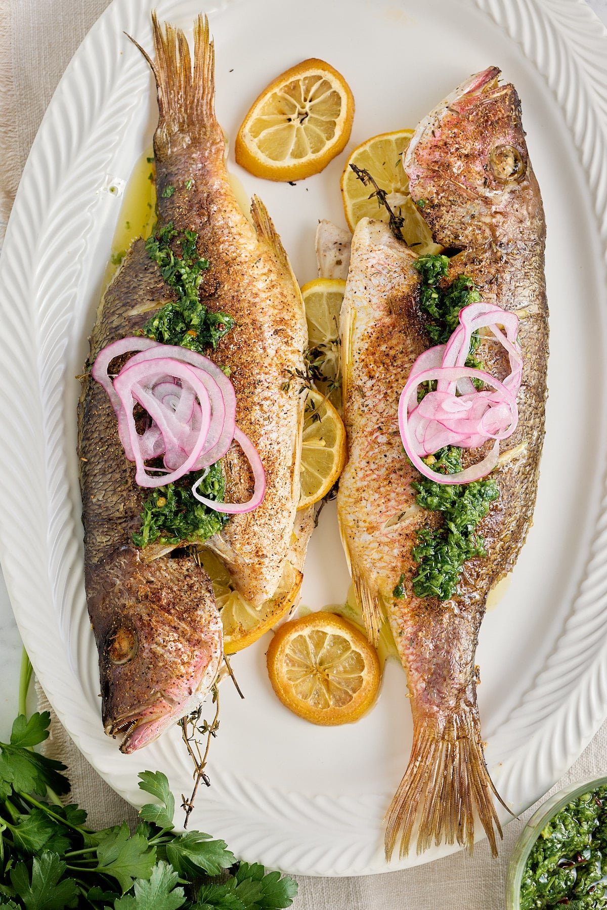 white serving platter with 2 cooked whole fish stuffed with fresh thyme and lemon slices and garnished with red onion and a drizzle of olive oil