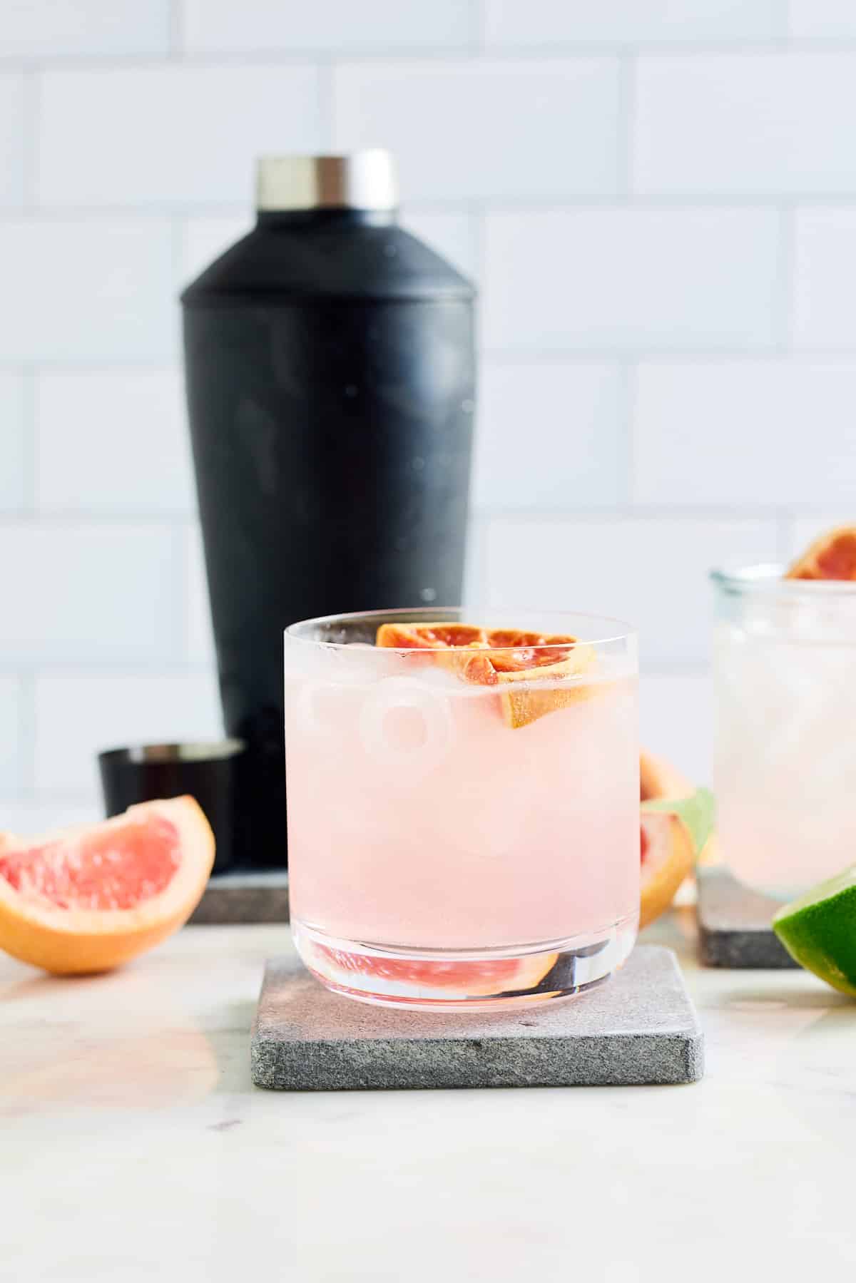 glass filled with pink cocktail topped with a slice of grapefruit and cocktail shaker with wedges of grapefruit and lime set alongside