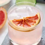 glass filled with pink cocktail topped with a slice of grapefruit and wedges of grapefruit and lime set alongside