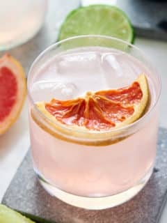 glass filled with pink cocktail topped with a slice of grapefruit and wedges of grapefruit and lime set alongside