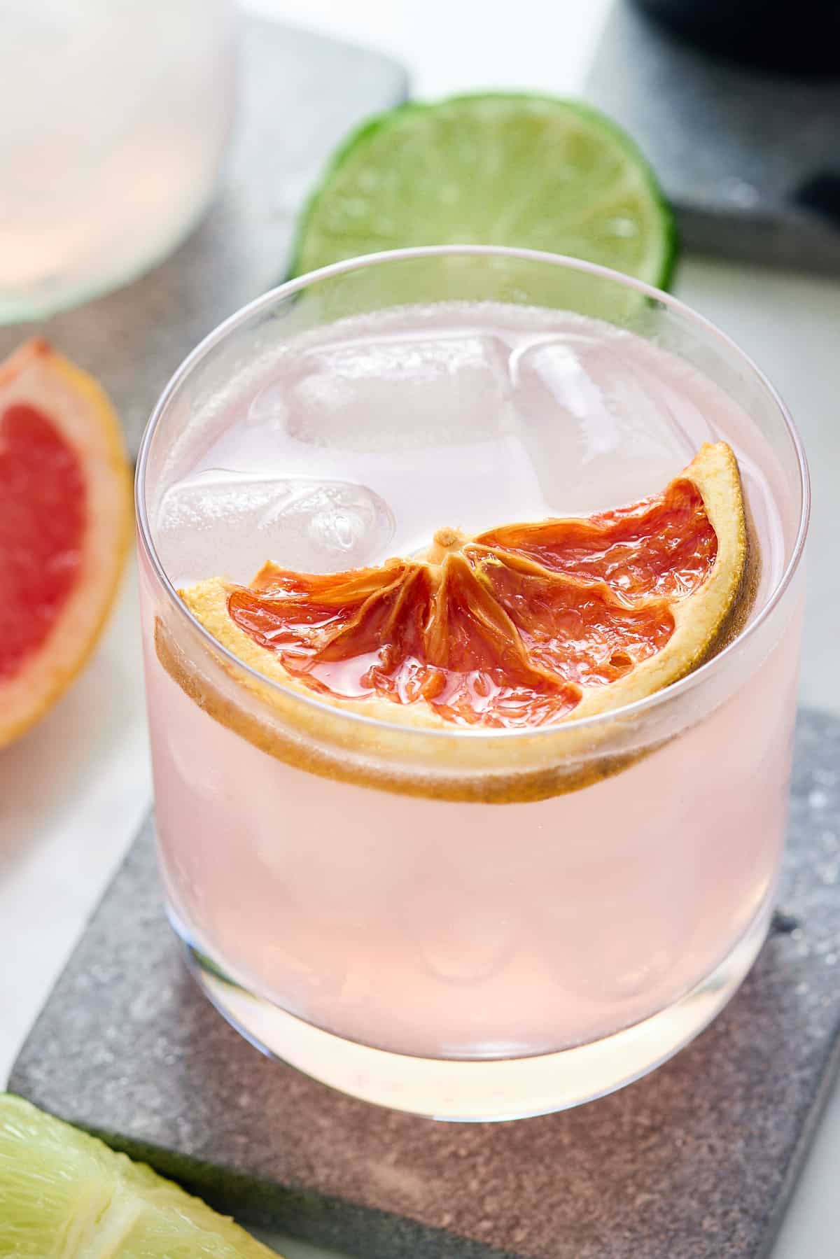 glass filled with pink cocktail topped with a slice of grapefruit and wedges of grapefruit and lime set alongside