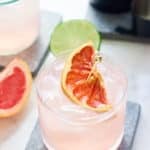 two glasses filled with pink cocktail topped with a slice of grapefruit and wedges of grapefruit and lime set alongside