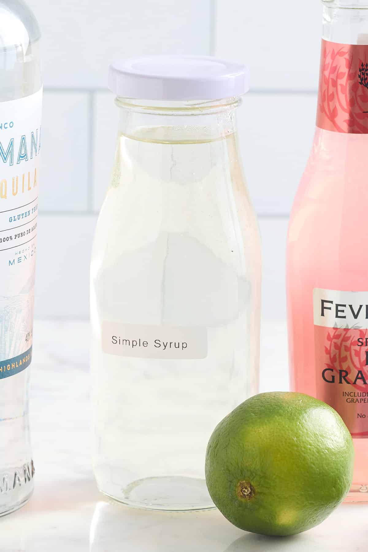 glass bottle filled with simple syrup with a bottle of tequila, grapefruit tonic and a fresh lime