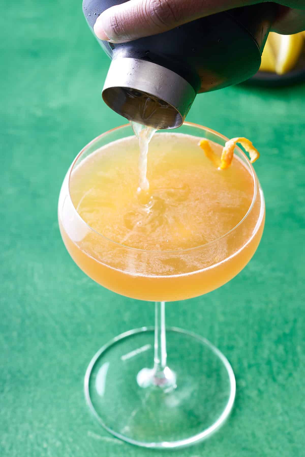 coupe glass filled with Hennessy sidecar cocktail being poured from a cocktail shaker and garnished with a strip of orange peel