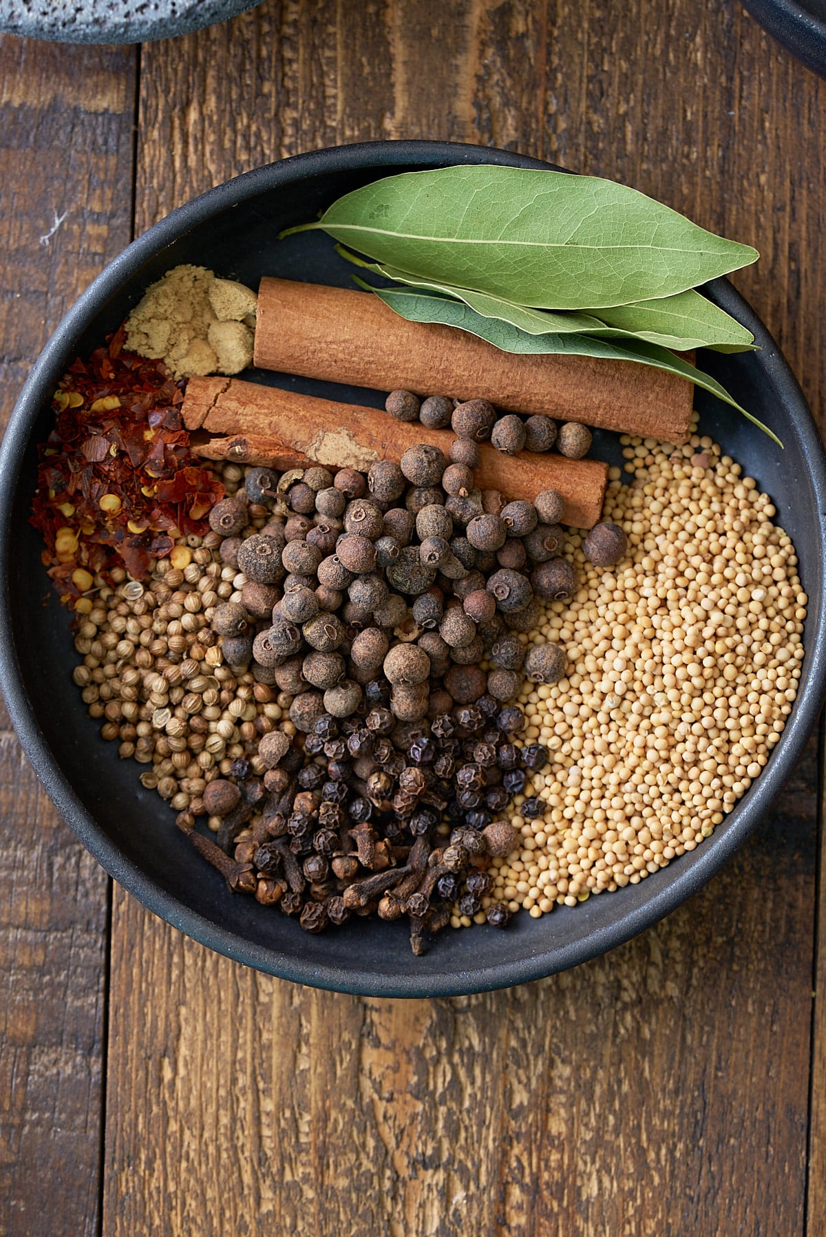 A black bowl filled with pickling spice recipe ingredients, including red pepper flakes, allspice berries, mustard seeds, black peppercorns, cinnamon sticks, bay leaves and coriander seeds.