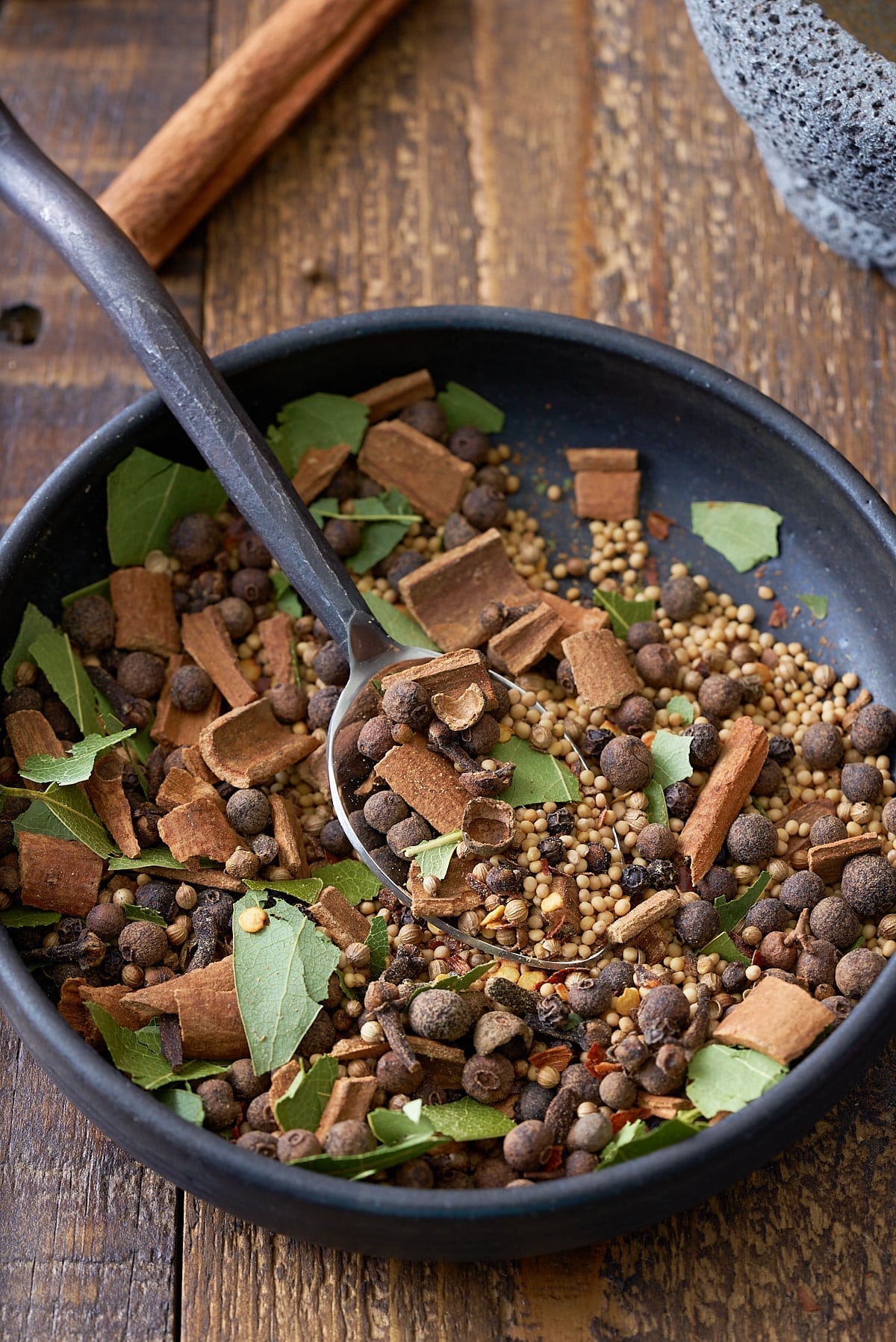 A black bowl filled with a combination of pickling spice, including dried whole spices including allspice berries, mustard seeds, black peppercorns, crushed cinnamon sticks, bay leaves and coriander seeds.