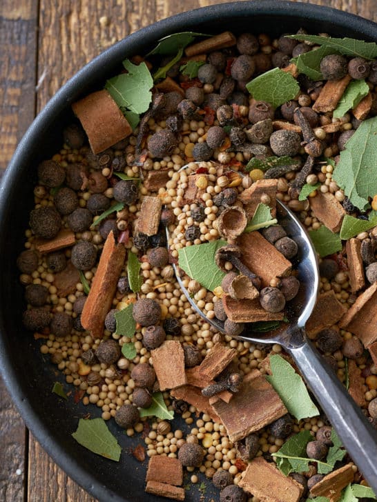 A black bowl filled with a combination of pickling spice, including dried whole spices including allspice berries, mustard seeds, black peppercorns, crushed cinnamon sticks, bay leaves and coriander seeds.