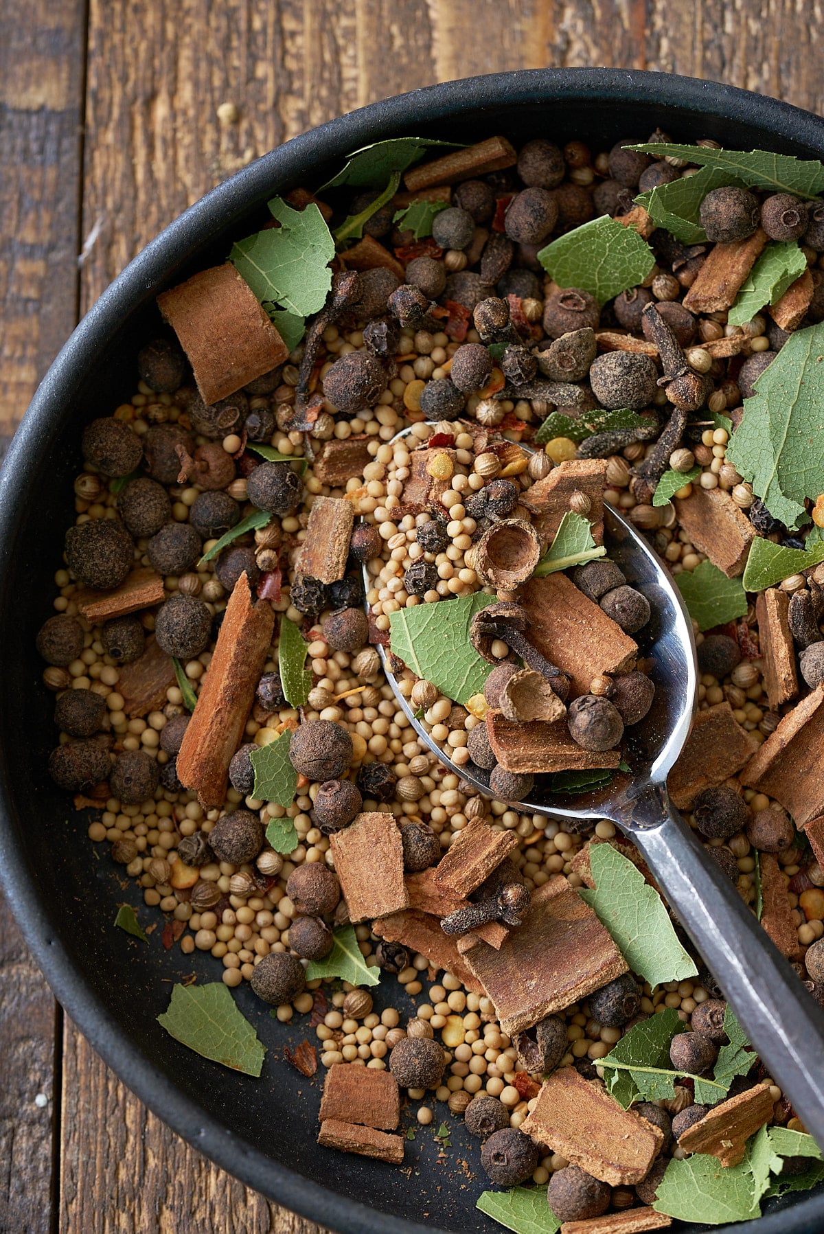 A black bowl filled with a combination of pickling spice, including dried whole spices including allspice berries, mustard seeds, black peppercorns, crushed cinnamon sticks, bay leaves and coriander seeds.