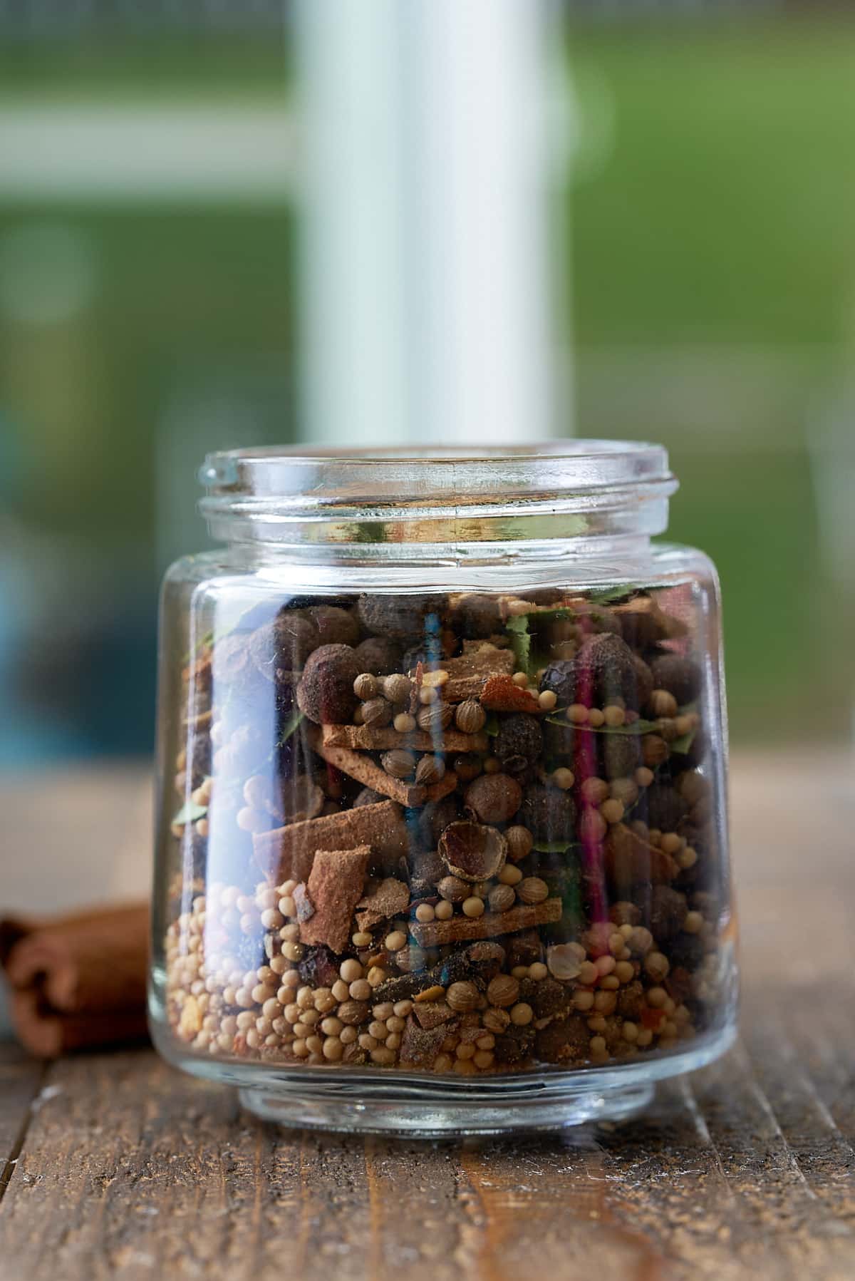 A glass jar filled with a variety of whole spices including allspice berries, mustard seeds, black peppercorns, cloves, crushed cinnamon sticks, bay leaves and coriander seeds, set on a wooden board.