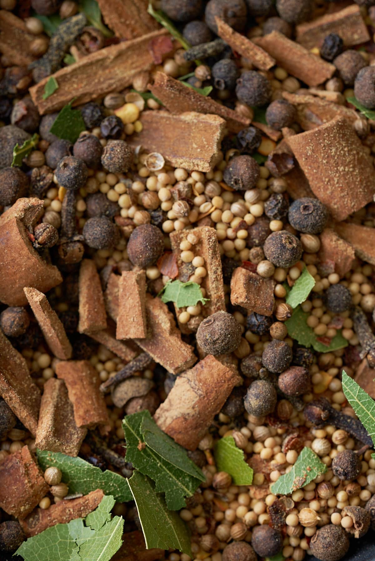 Whole spices including allspice berries, mustard seeds, black peppercorns, cloves, crushed cinnamon sticks, bay leaves and coriander seeds, set on a wooden board.