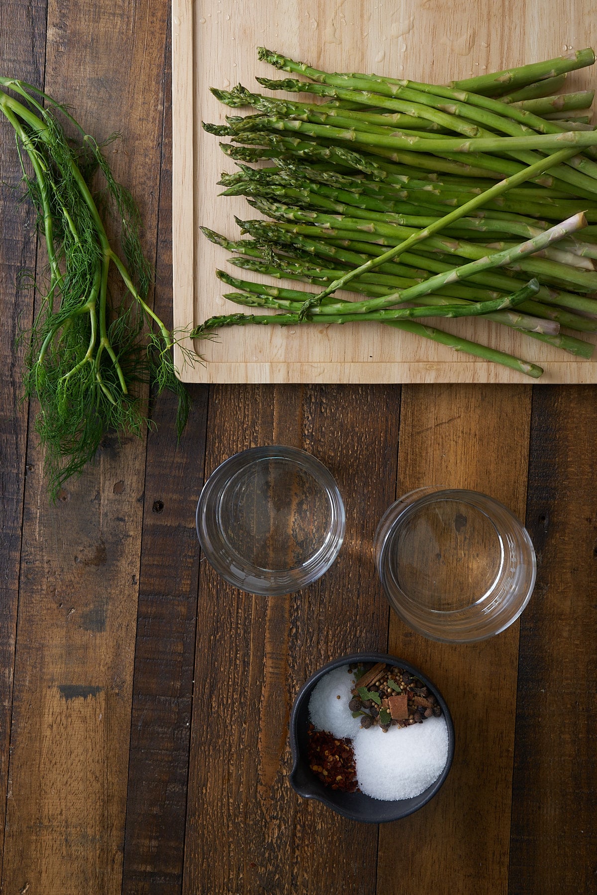 Pickled asparagus recipe ingredients including asparagus spears set on a wooden board, fresh sprigs of dill, water, vinegar and a bowl with pickling spices, salt and sugar.