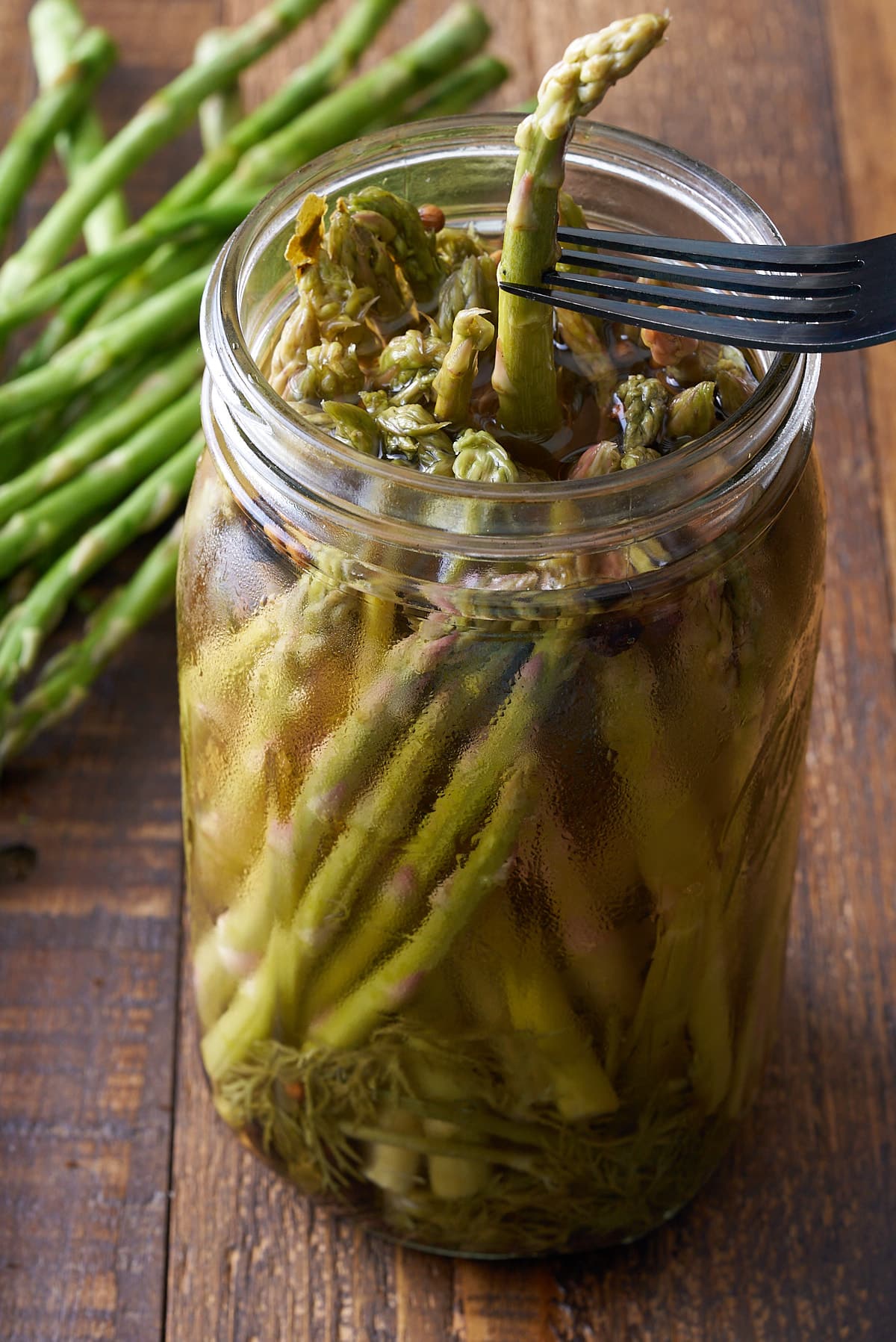 A glass mason jar filled with pickled asparagus and a fork removing an asparagus spear.