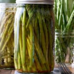 Two glass mason jars filled with pickled asparagus with a jar of fresh asparagus set alongside.
