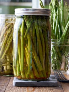 Two glass mason jars filled with pickled asparagus with a jar of fresh asparagus set alongside.