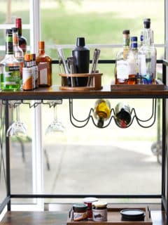 photo of bar cart in front of window filled with home bar essentials, like cocktail shakers, liquors, and liquers