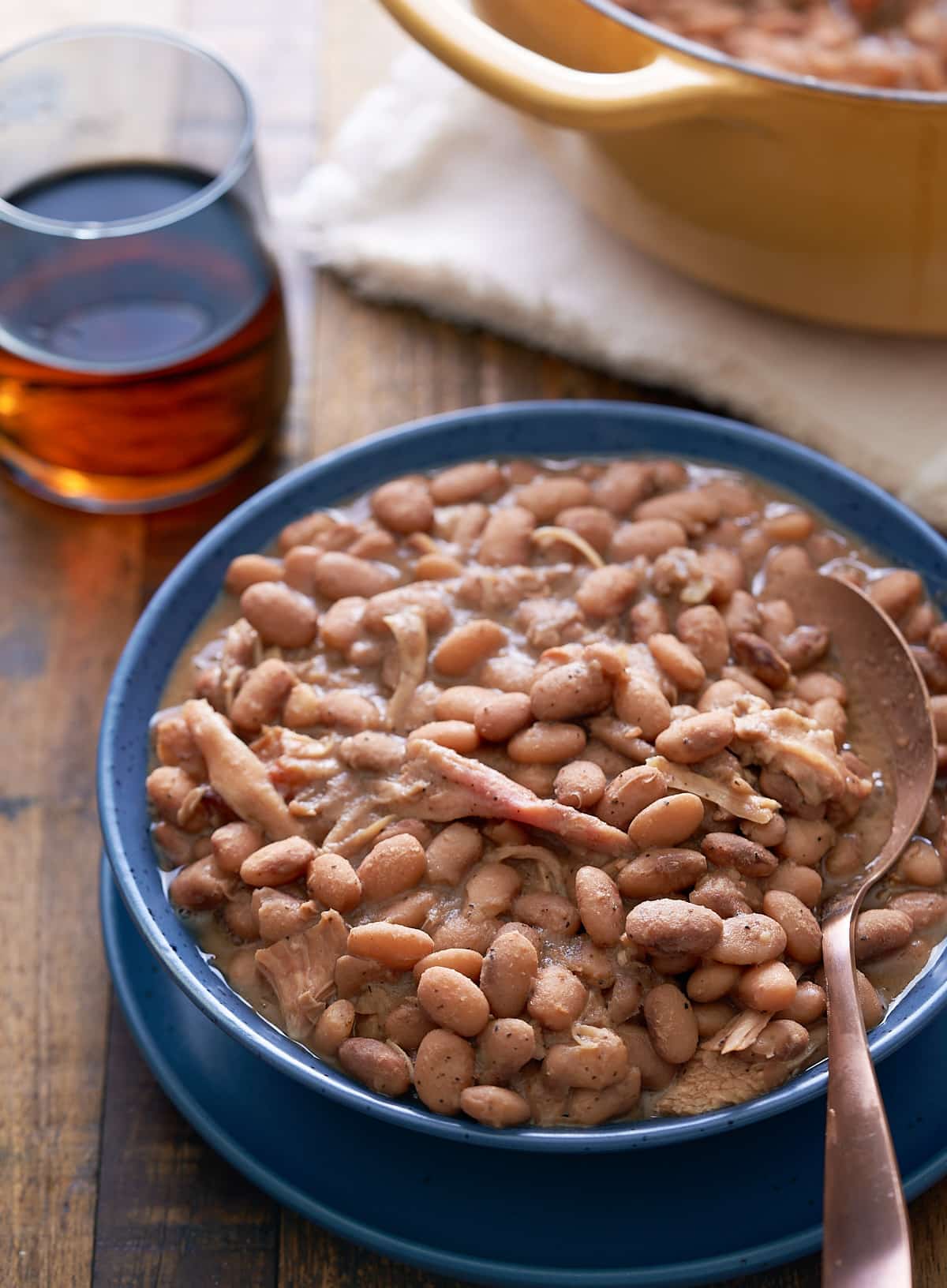 A blue bowl and spoon filled with southern style pinto beans with shredded turkey leg, with a Dutch oven and a glass of cola set alongside.