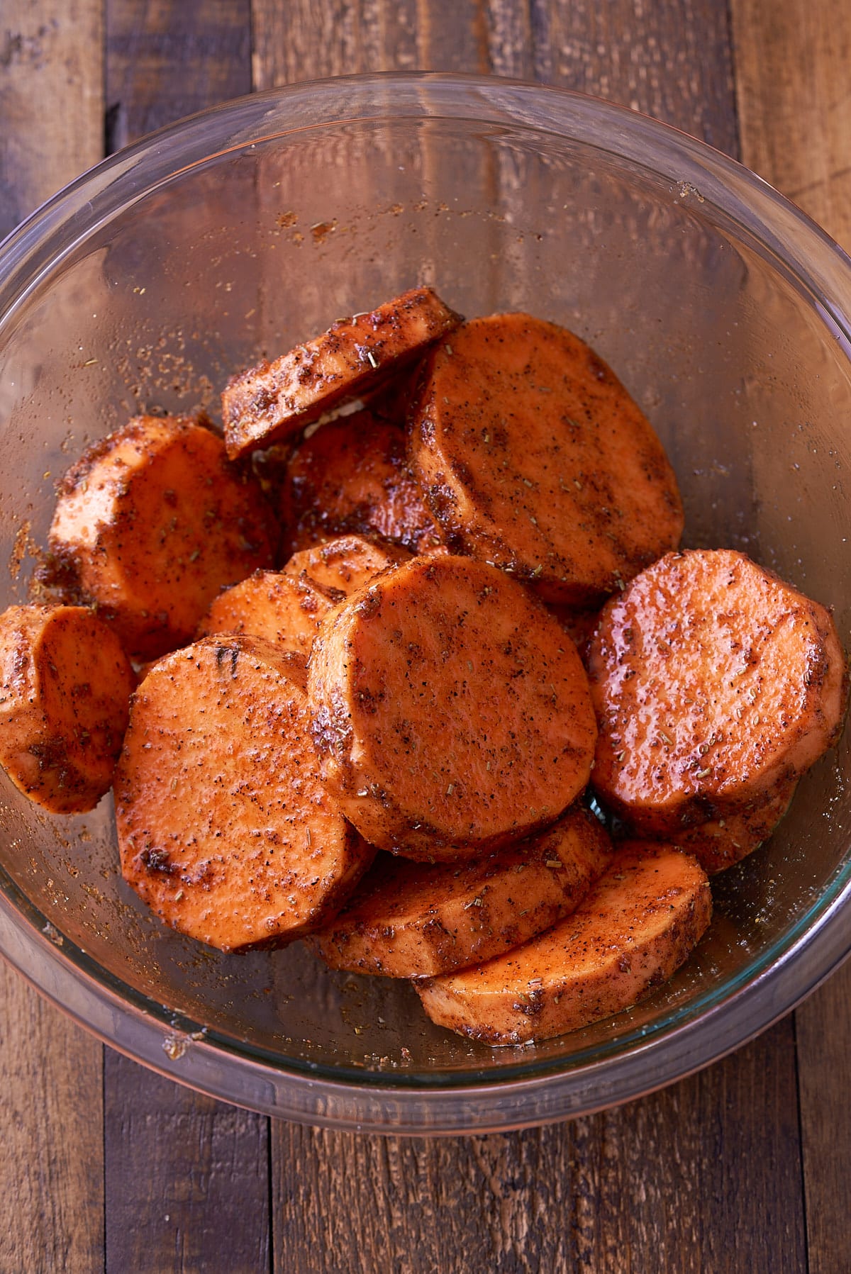 A glass bowl filled with sweet potato rounds covered in oil, spices, salt and sugar.