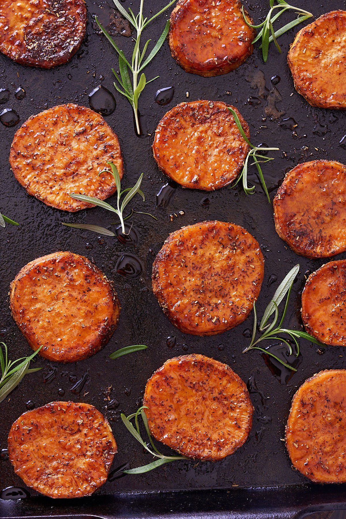 Close up image of baked sweet potato rounds on a baking sheet with springs of fresh rosemary.