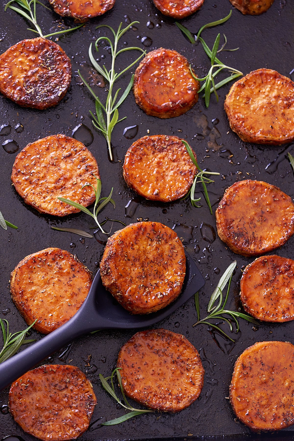 Close up image of baked sweet potato rounds on a baking sheet with springs of fresh rosemary.