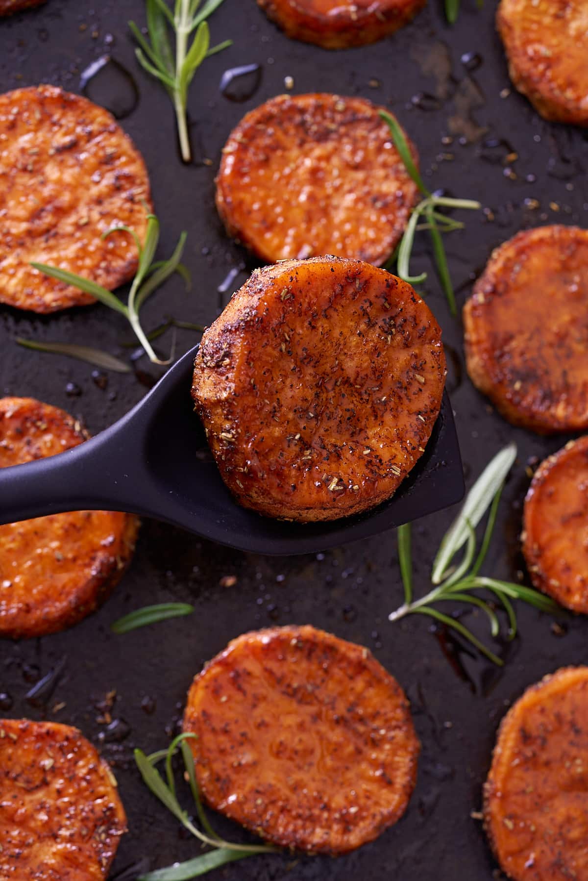 Close up image of baked sweet potato rounds on a baking sheet with springs of fresh rosemary.