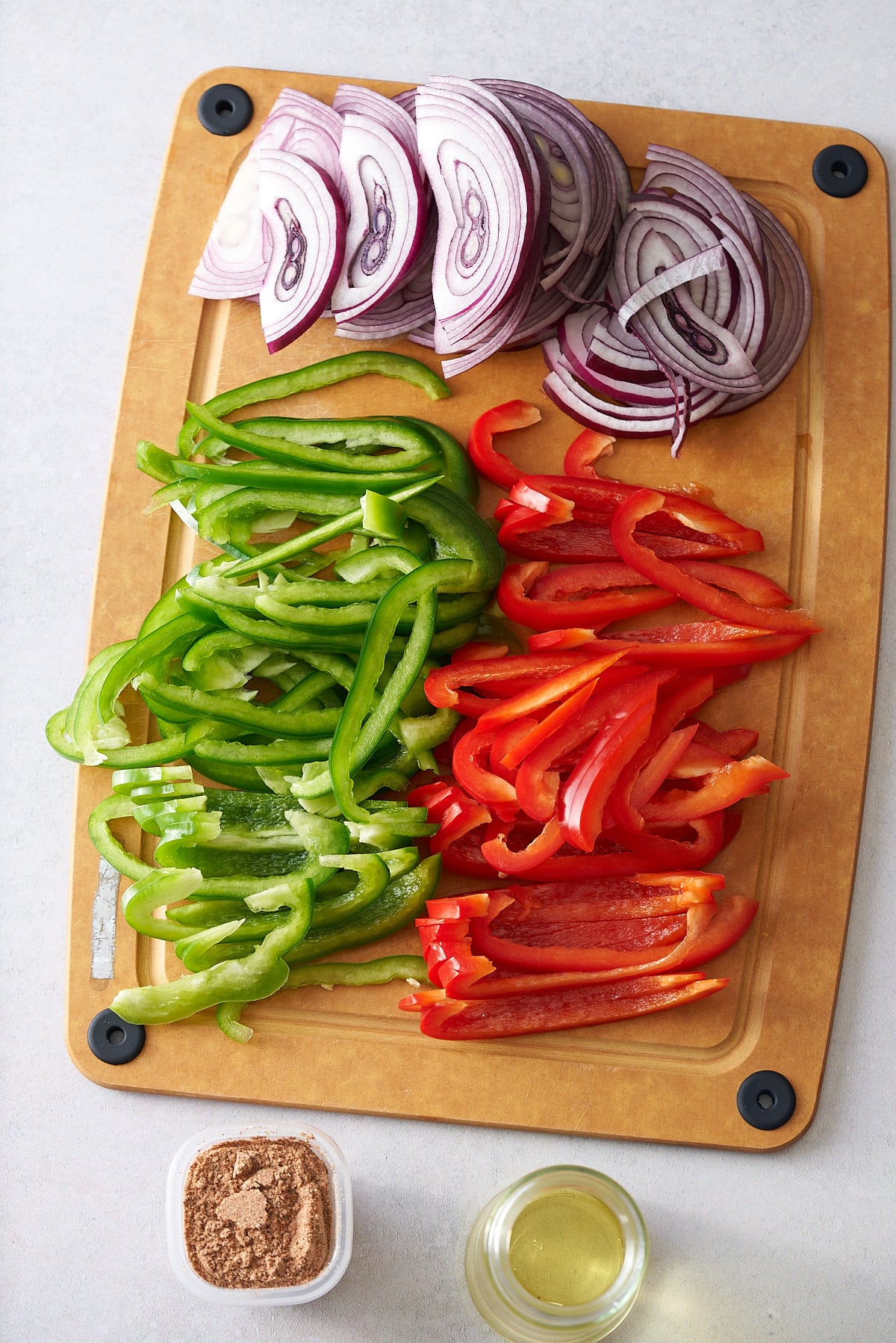 Sliced red onion and red and green bell peppers on a wooden chopping board, with pots of fajita seasoning and olive oil set alongside.