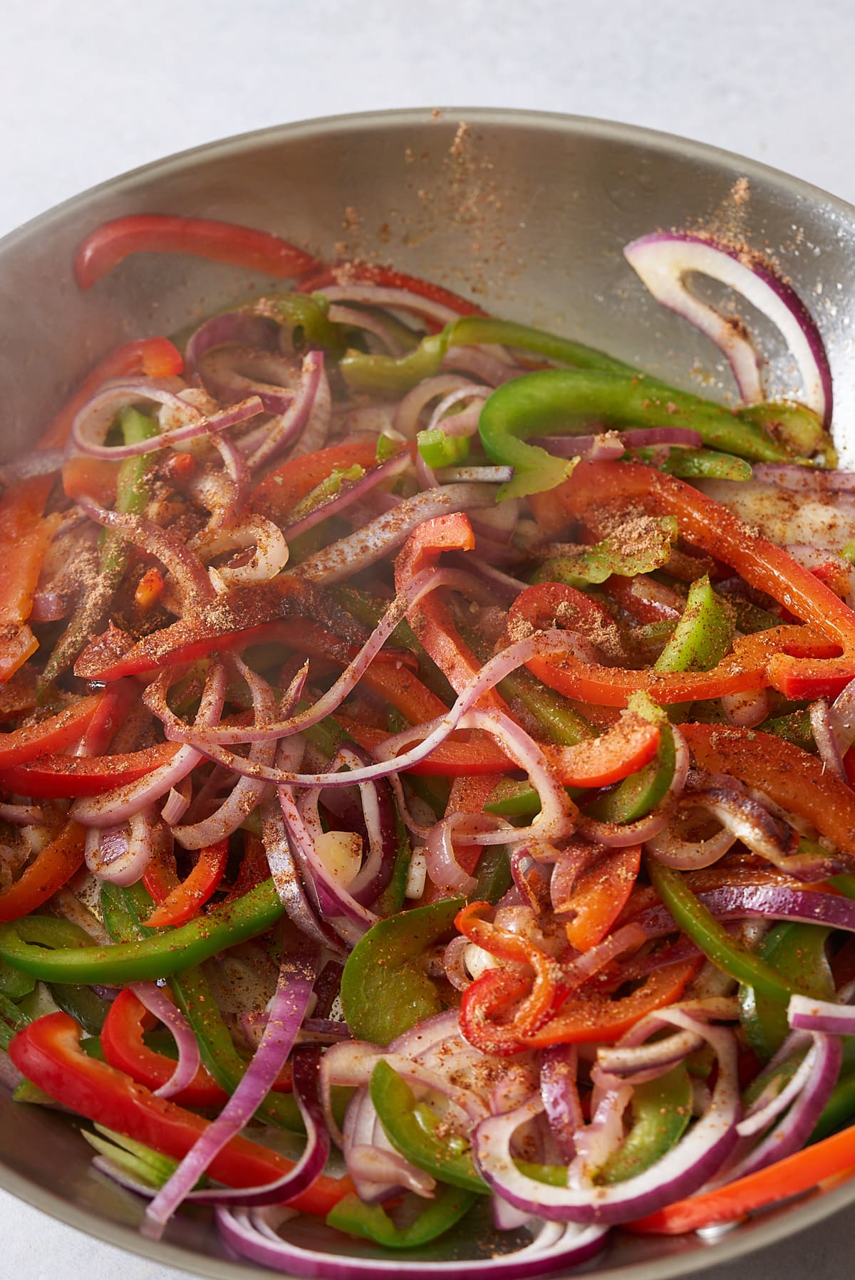 Sliced red onion and red and green bell peppers coated in fajitas seasoning, cooking in a large frying pan.