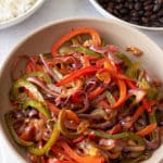 A large serving dish filled with fajitas vegetables, with bowls of steamed white rice and black beans set alongside.