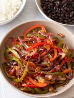 A large serving dish filled with fajitas vegetables, with bowls of steamed white rice and black beans set alongside.
