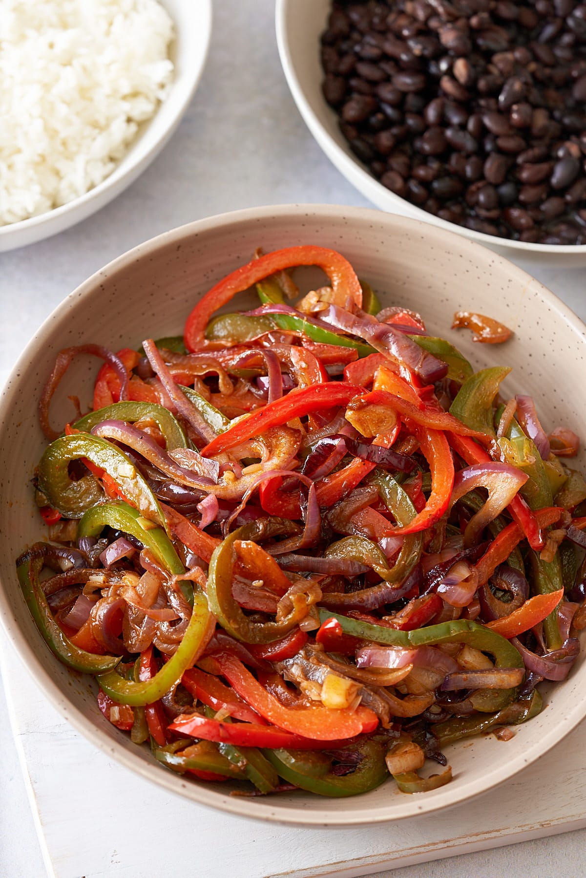 A large serving dish filled with fajitas vegetables, with bowls of steamed white rice and black beans set alongside.