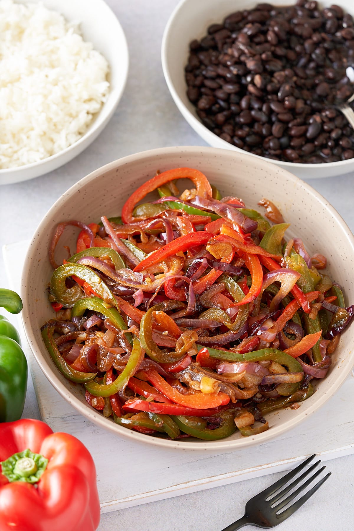 A large serving dish filled with fajitas vegetables, with a red and green bell peppers, and bowls of steamed white rice and black beans set alongside.