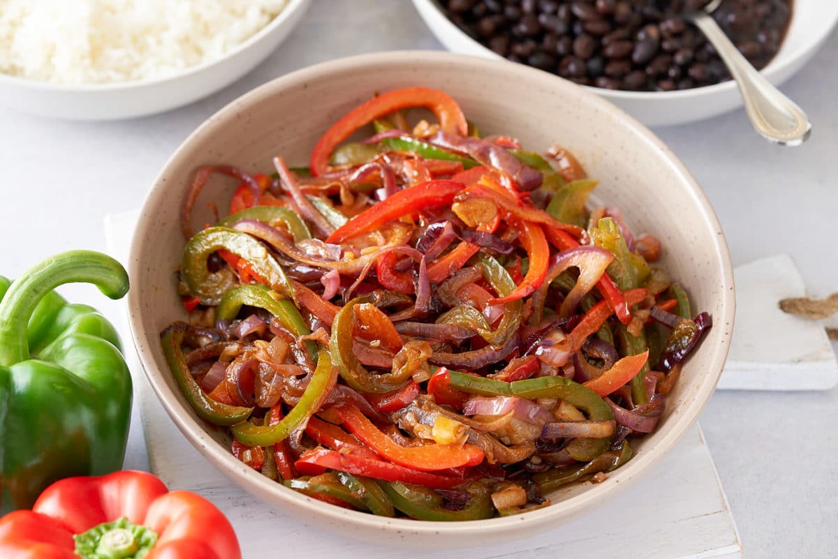 A large serving dish filled with fajitas vegetables, with a red and green bell peppers, and bowls of steamed white rice and black beans set alongside.